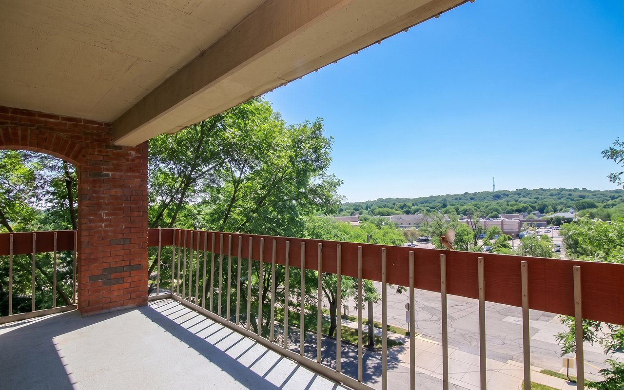 Covered balcony overlooking leafy trees, low-rise buildings, and a wooded hill.