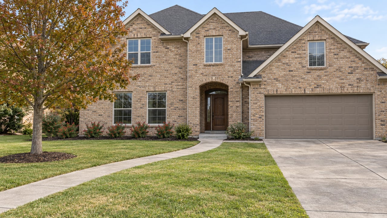 Buff-brick house with pitched charcoal roofs, a broad front lawn, a curved concrete walk, and an oak beginning to turn russet.