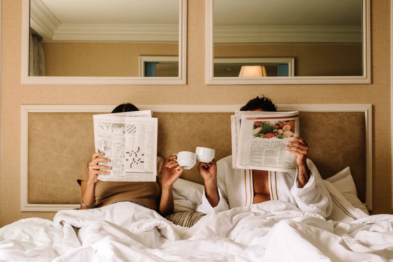 A couple reads newspapers in bed, holding coffee cups beneath white covers.