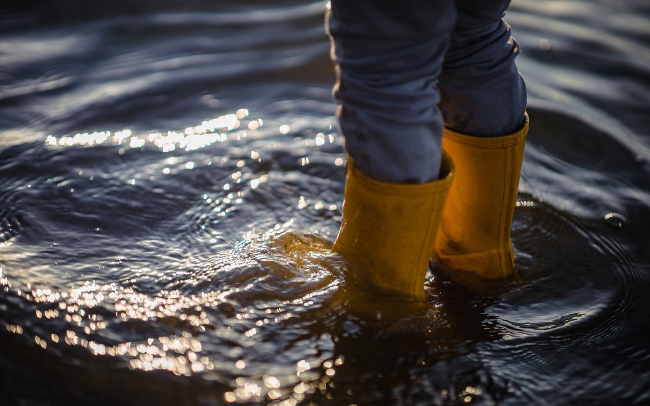 Person in yellow rubber boots standing in sunlit floodwater.