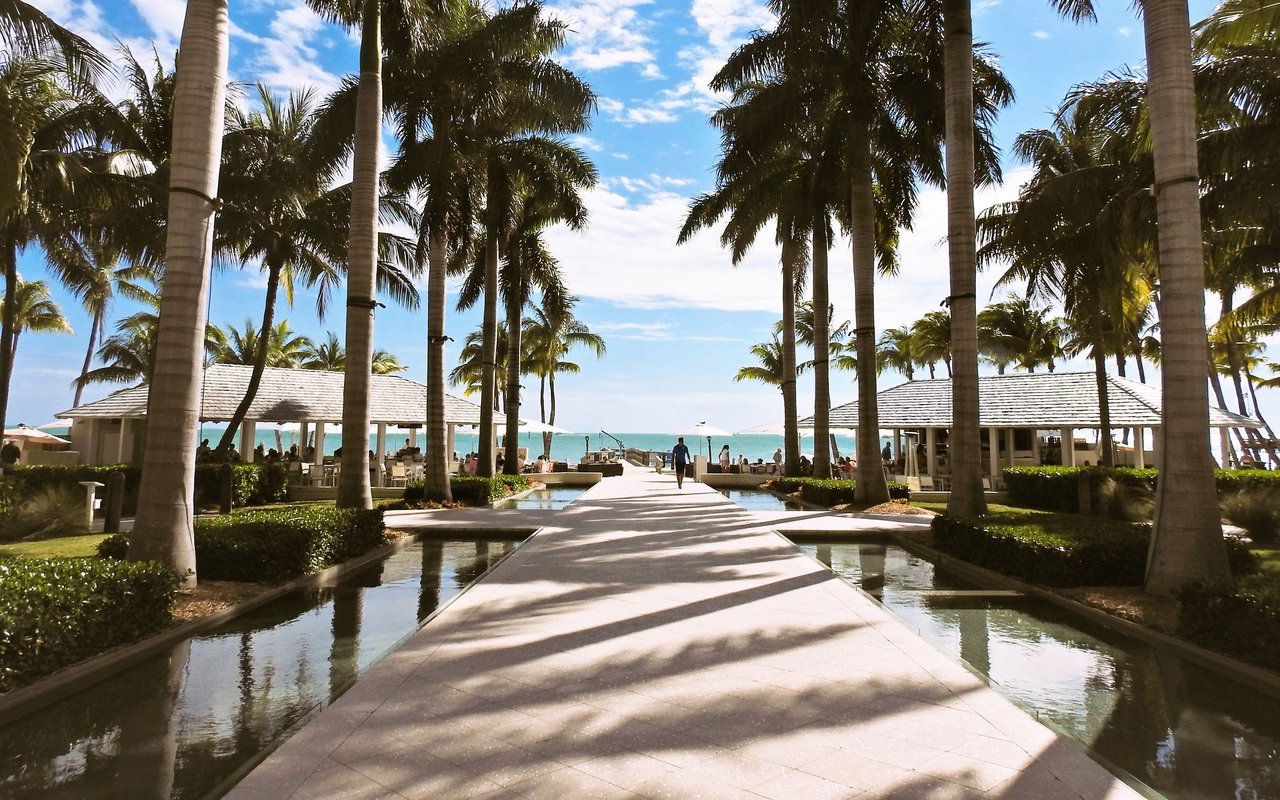 Palm-lined resort walkway and reflecting pools lead toward open-air pavilions and the ocean.
