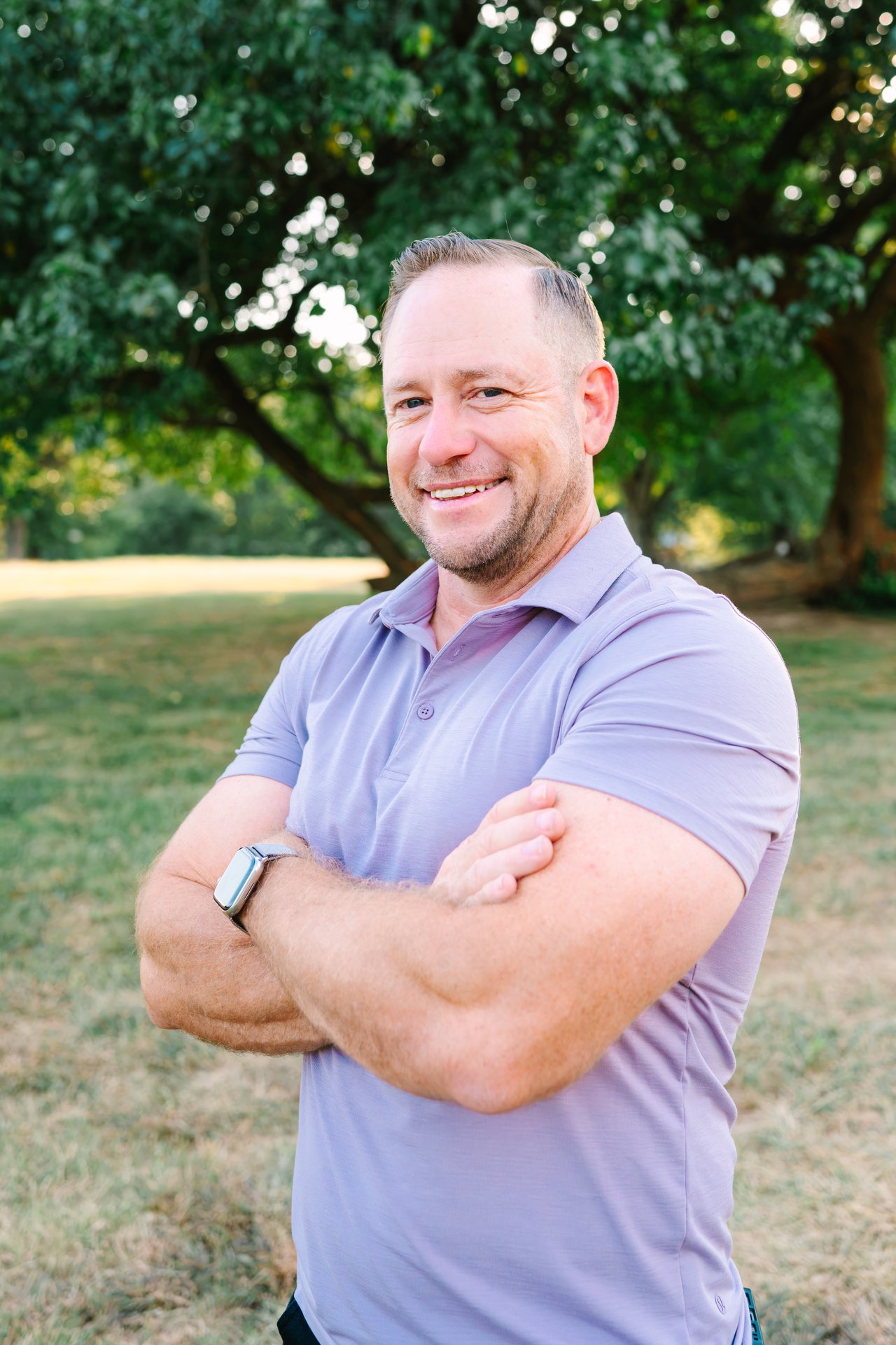Joshua Ratliff, smiling with arms crossed outdoors beneath leafy trees.