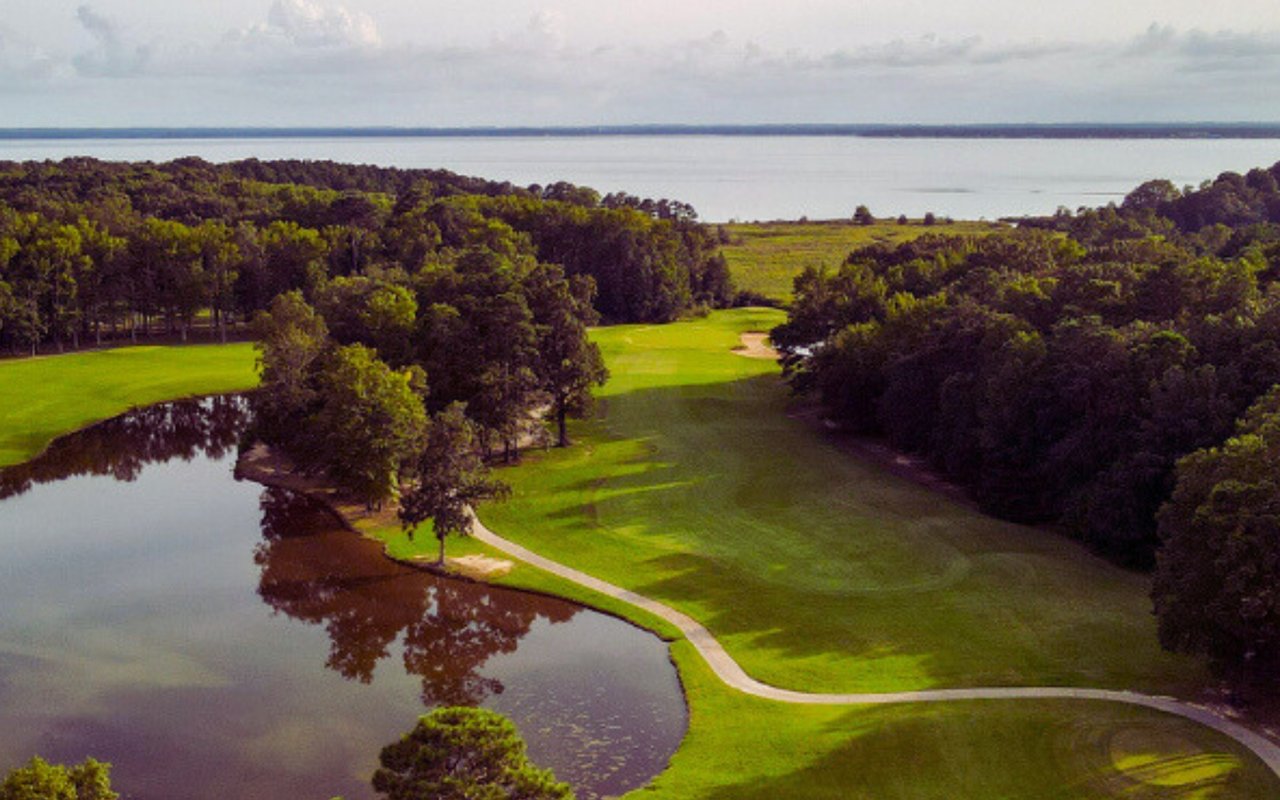 Aerial golf course with tree-lined fairways, a pond, and water in the distance.