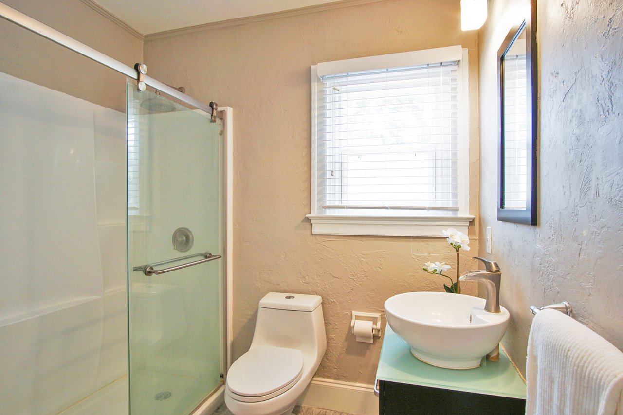 Bright modern bathroom with textured beige walls, featuring a glass-enclosed shower, sleek toilet, and a round vessel sink. Sunlight streams through a window.