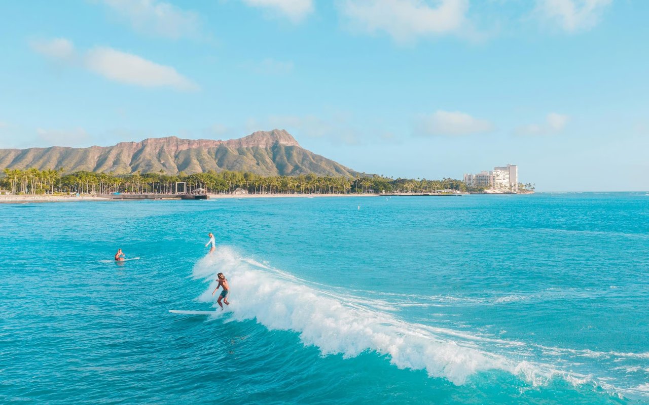 Surfers ride a wave off Waikiki Beach, with Diamond Head and palm-lined shore in the distance.