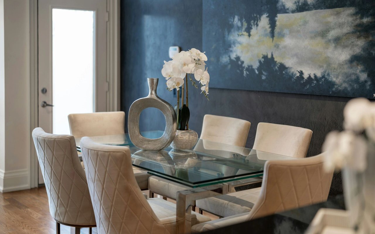 Dining room with a glass table, cream chairs, white orchids, and a large blue abstract painting.
