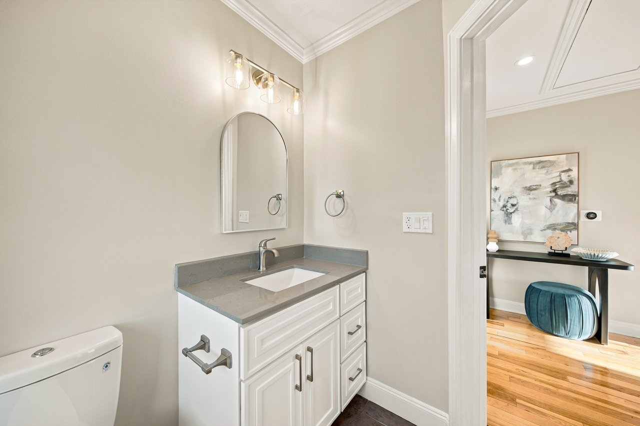 Bathroom with a white paneled vanity, gray countertop, arched mirror, and doorway to a furnished room.