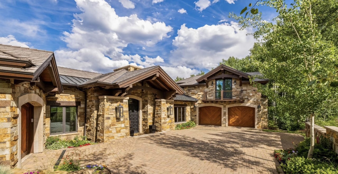 Stone-clad home with timber gables, two wooden garage doors, and a landscaped paved courtyard.