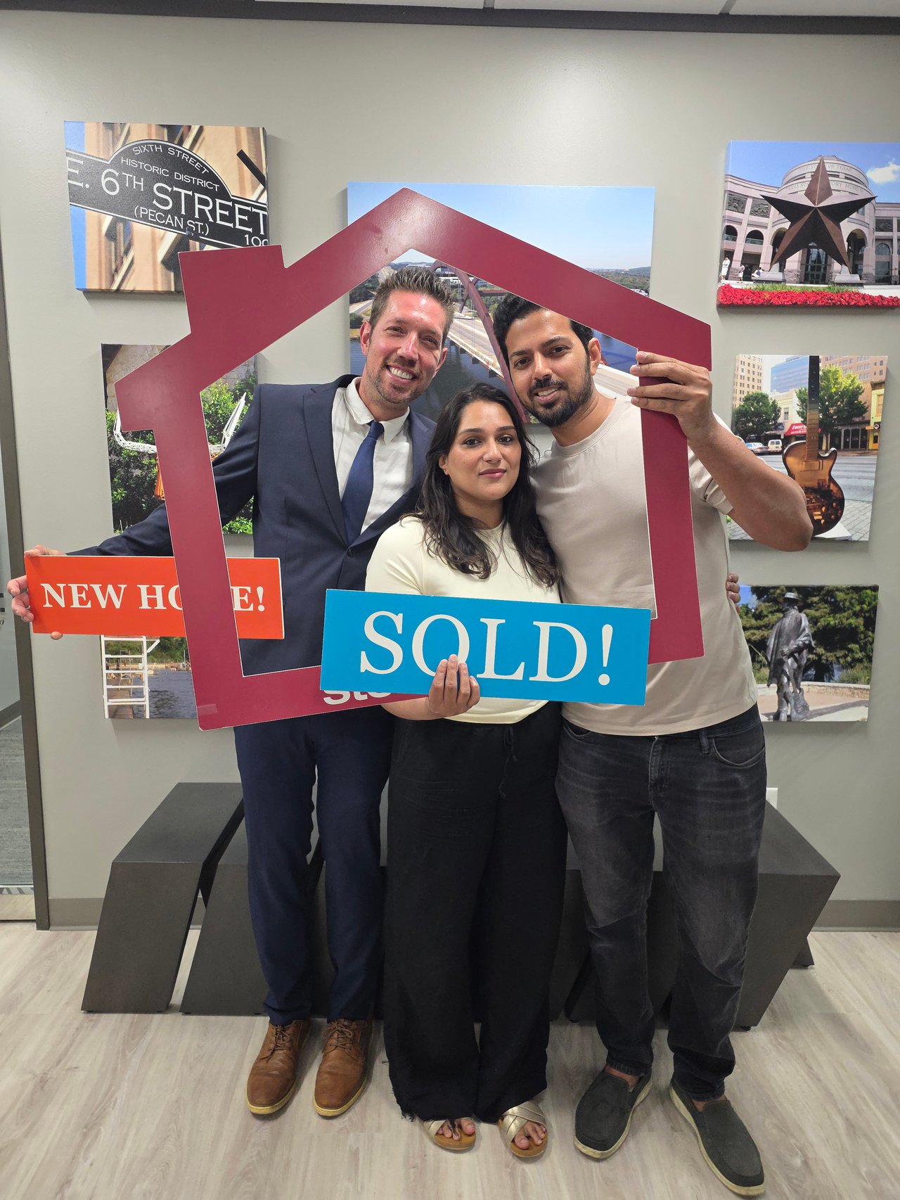 Three adults pose with “SOLD!” and “NEW HOME!” signs inside a real estate office.