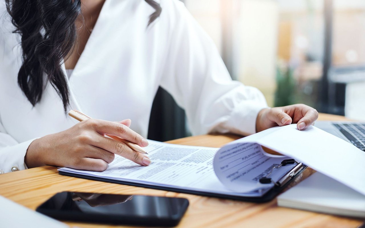 Woman reviewing paperwork on a clipboard beside a laptop and smartphone.