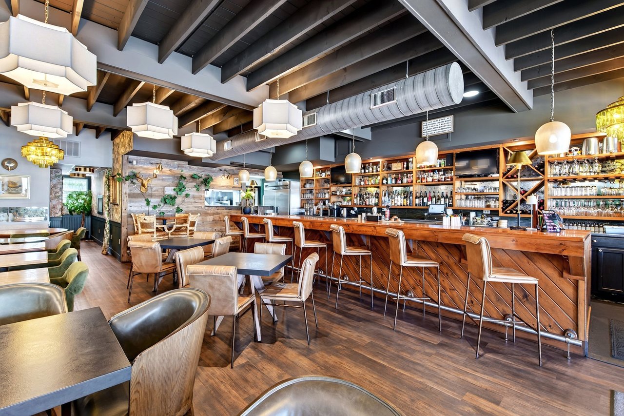 Restaurant dining room with a long wood bar, exposed beams, and tables and chairs.