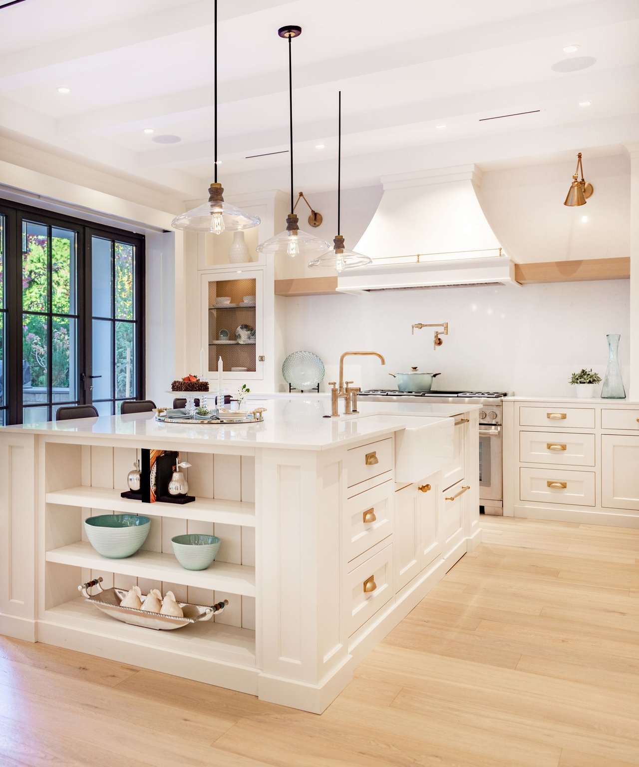 Bright white kitchen with a display-shelf island, brass fixtures, glass pendants, and black-framed garden doors.