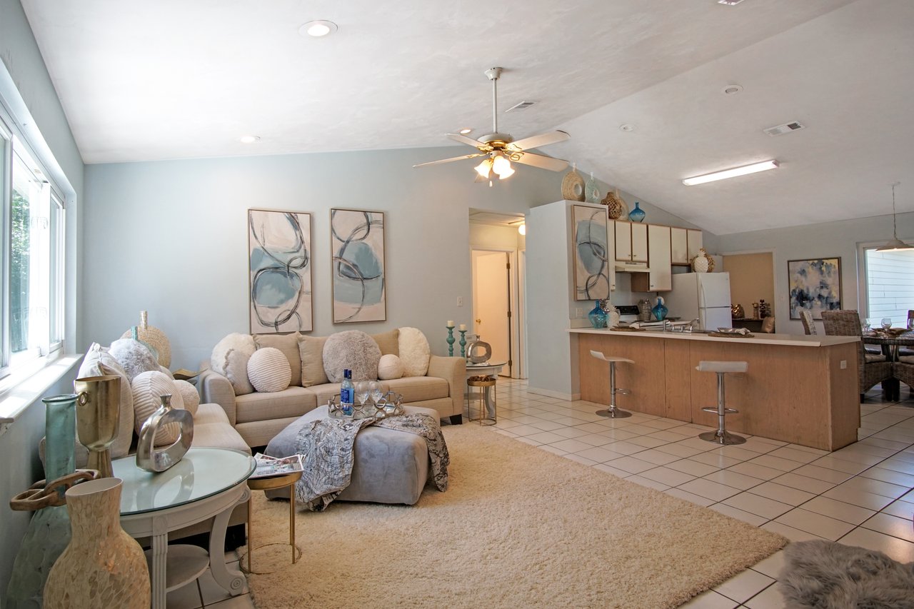 Spacious living room with light blue walls, abstract art, and a cozy beige couch adorned with round cushions. The bright kitchen features a wooden island with bar stools. The room is well-lit by natural light from large windows, creating a calm, inviting atmosphere.