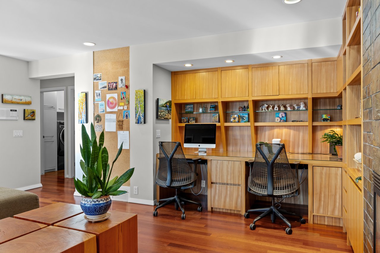 Home office with built-in wood desk and cabinetry, two black chairs, and a potted plant.