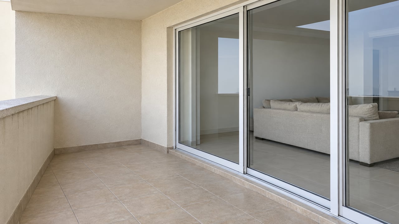 A cream stucco condominium balcony with sand-colored tile, white-framed sliding glass doors and a softly furnished living room beyond.