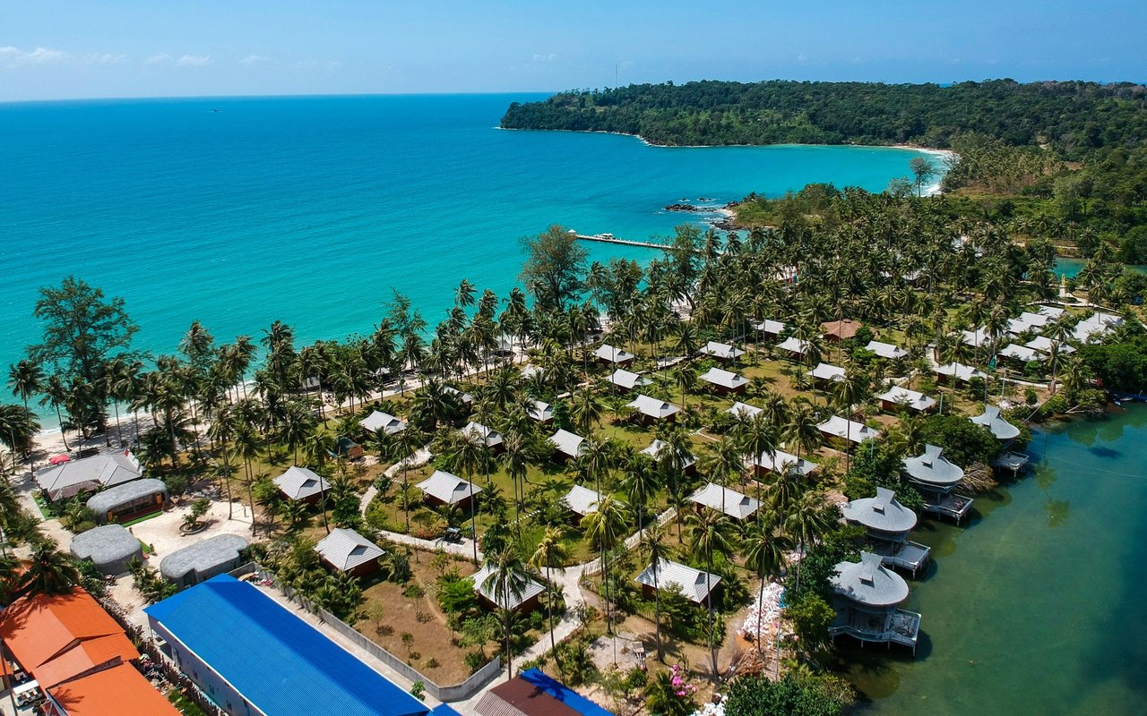 Aerial view of palm-covered coastal buildings beside a turquoise bay and curving shoreline.