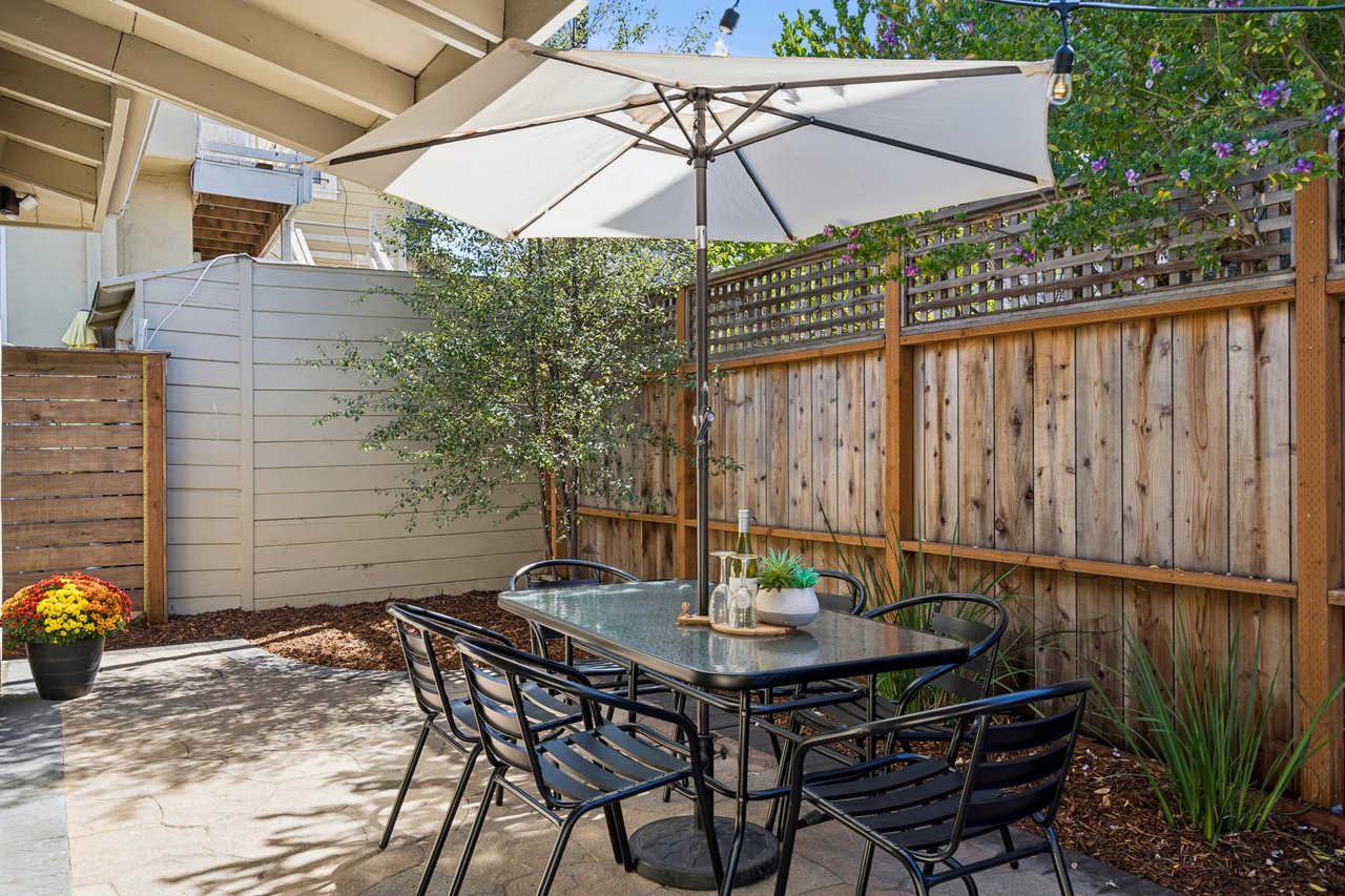 Outdoor dining patio beneath a large white umbrella, enclosed by a wooden fence and flowering vines.