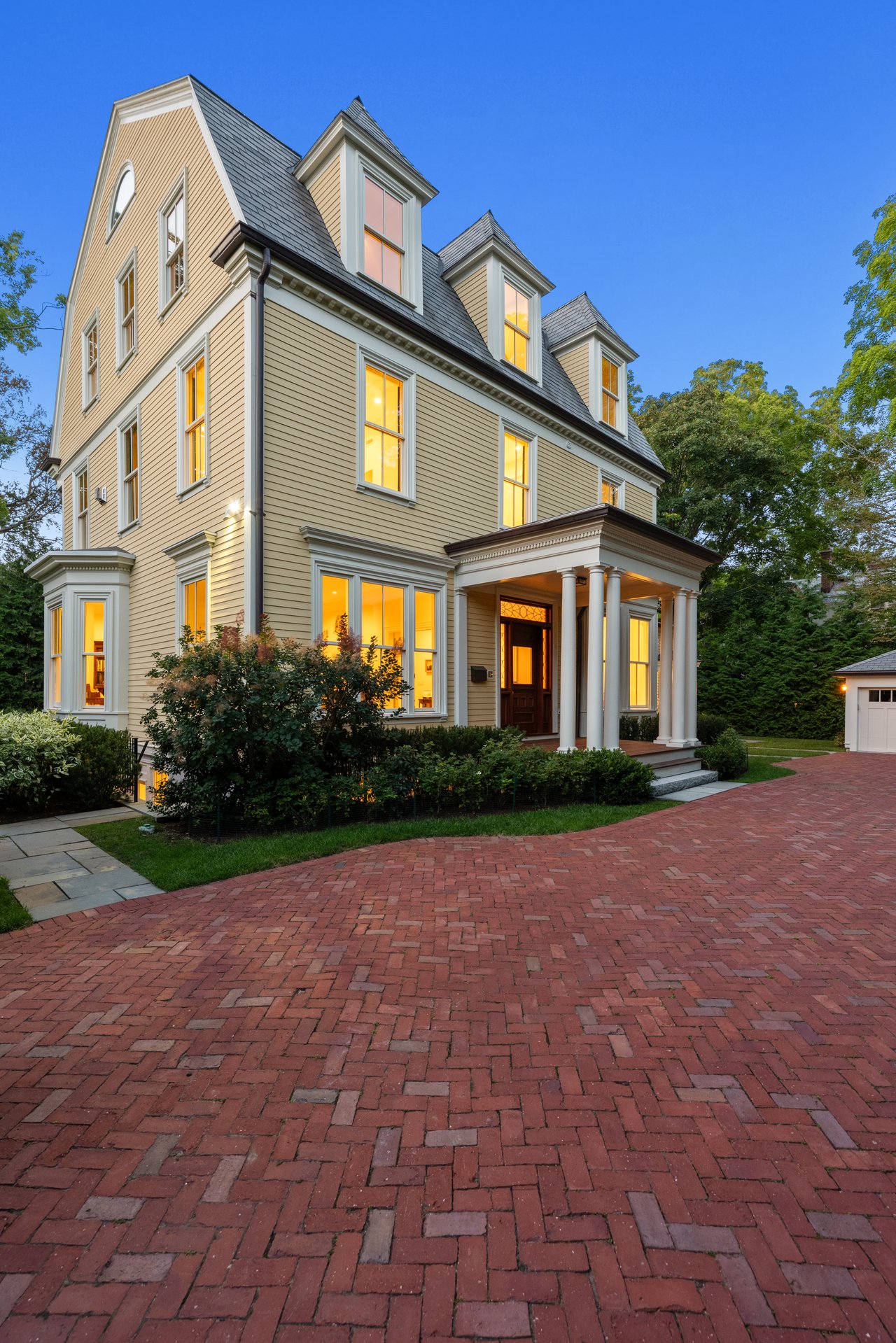 Victorian home with illuminated windows, steep slate roof, columned porch, and brick driveway.
