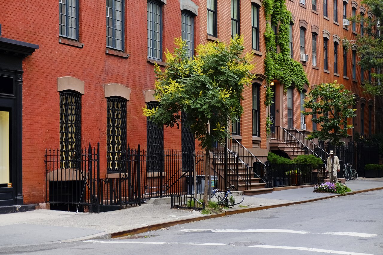 Red-brick urban residences with iron-railed stoops, leafy vines, and trees along the sidewalk.