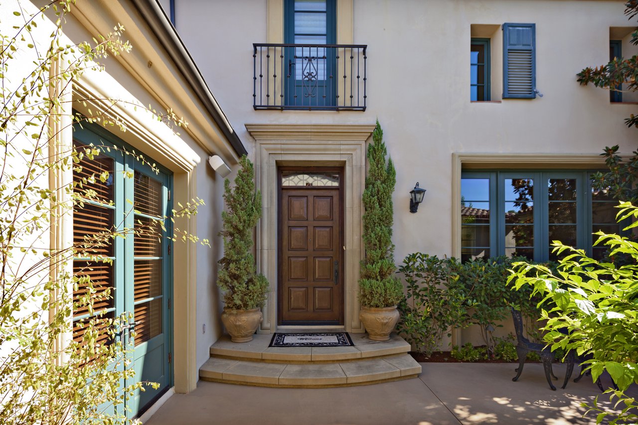 Home entrance with a paneled wood door, curved stone steps, potted evergreens, and a wrought-iron balcony.