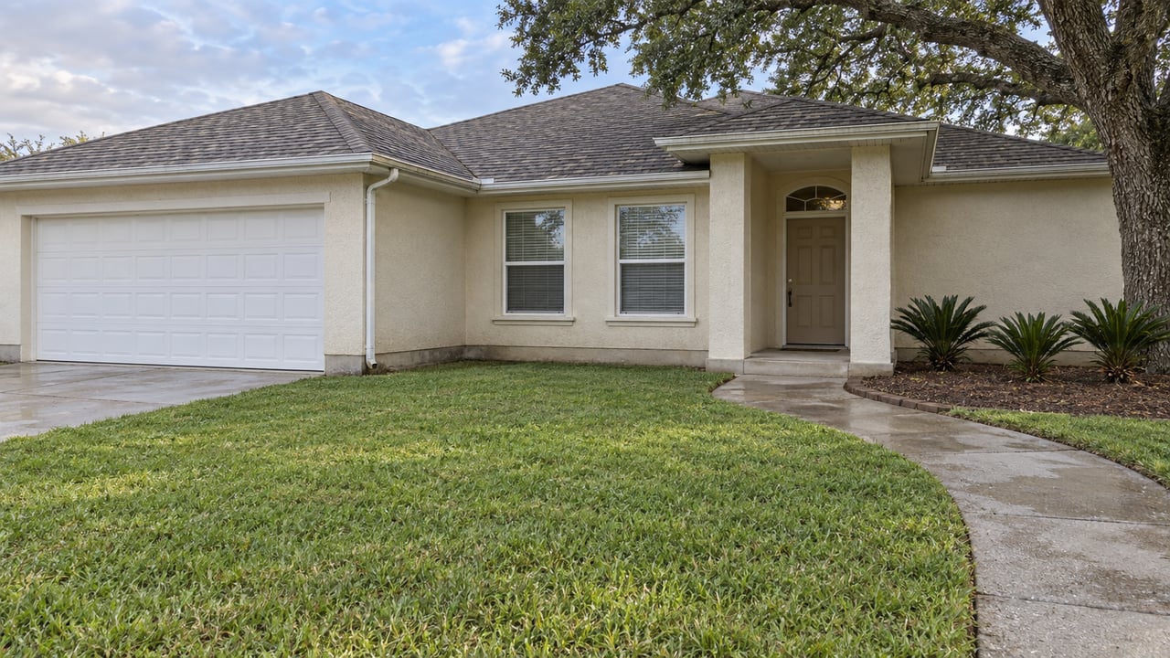 An ivory stucco single-story home with a broad lawn, recessed entry and spreading oak after rain.