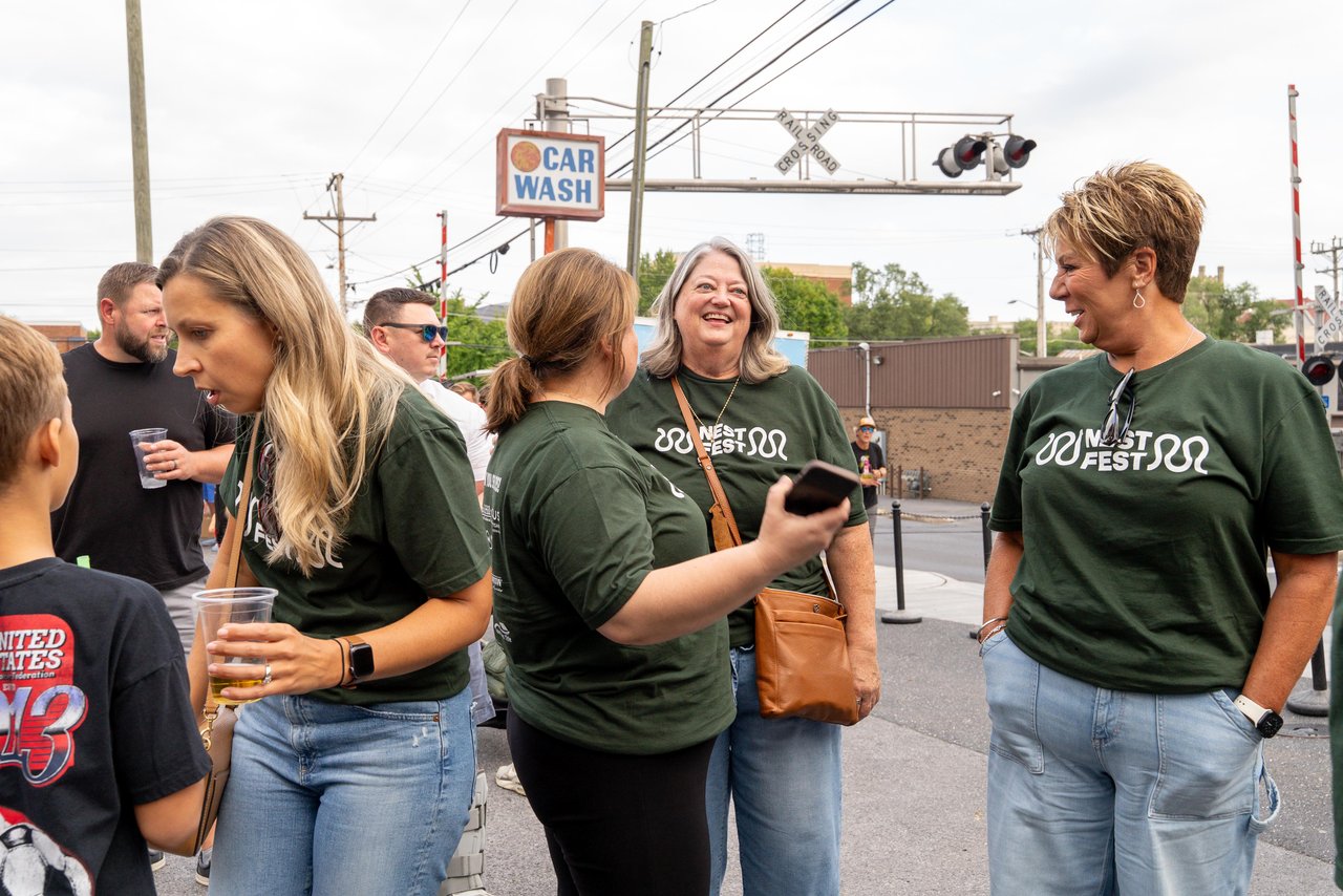 A group in green Nest Fest shirts chat outdoors near a railroad crossing.