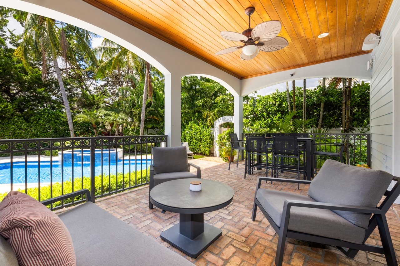 Covered brick patio with cushioned seating, a wooden ceiling, and a swimming pool surrounded by tropical landscaping.