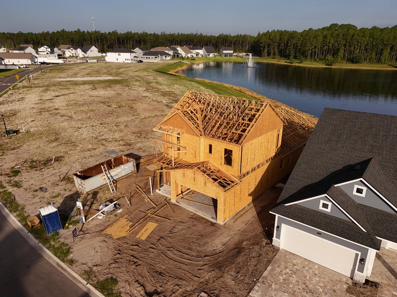 Aerial view of a wood-framed house under construction beside a pond and neighboring homes.