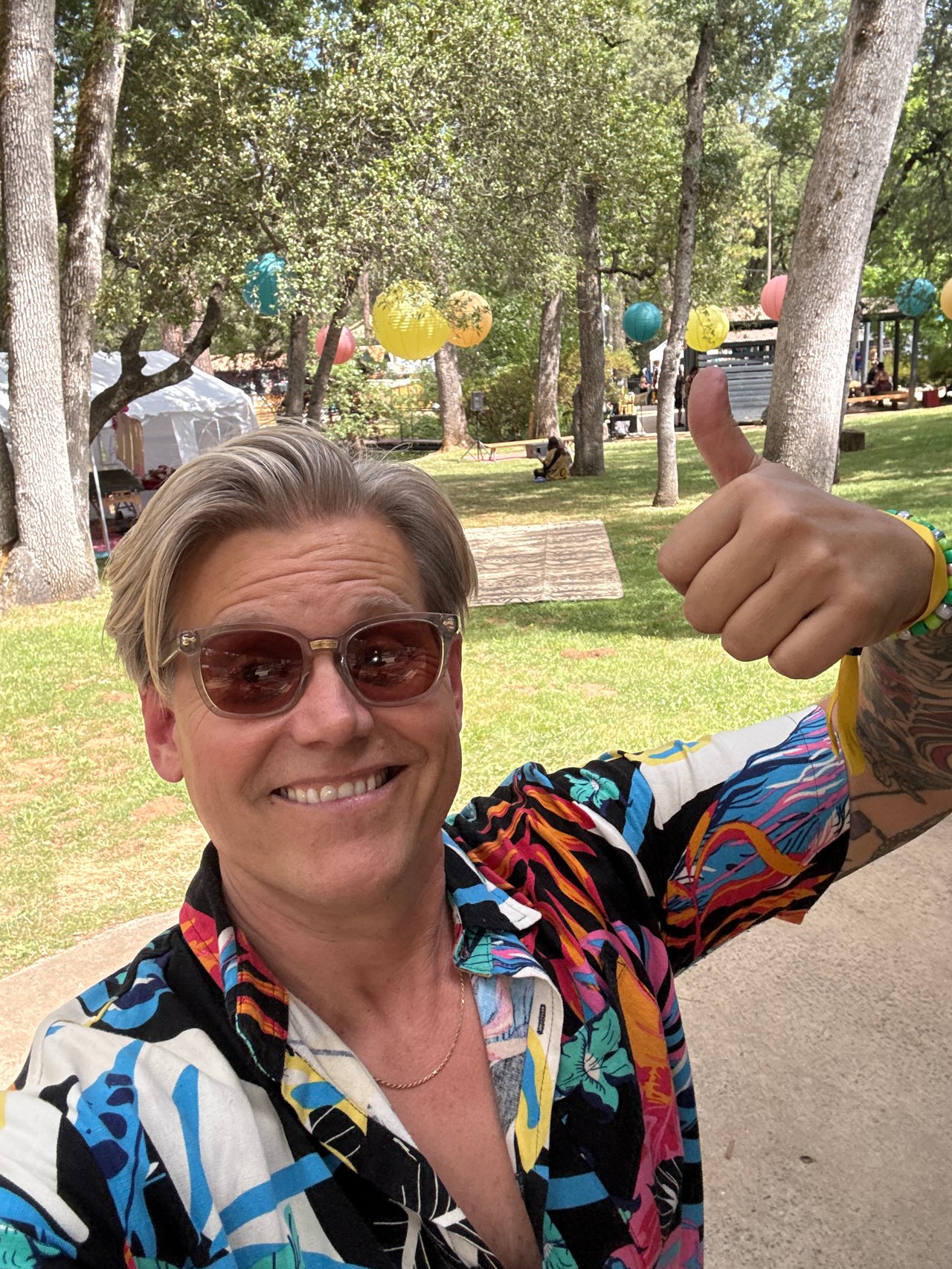Man in sunglasses and a colorful shirt gives a thumbs-up at an outdoor gathering.