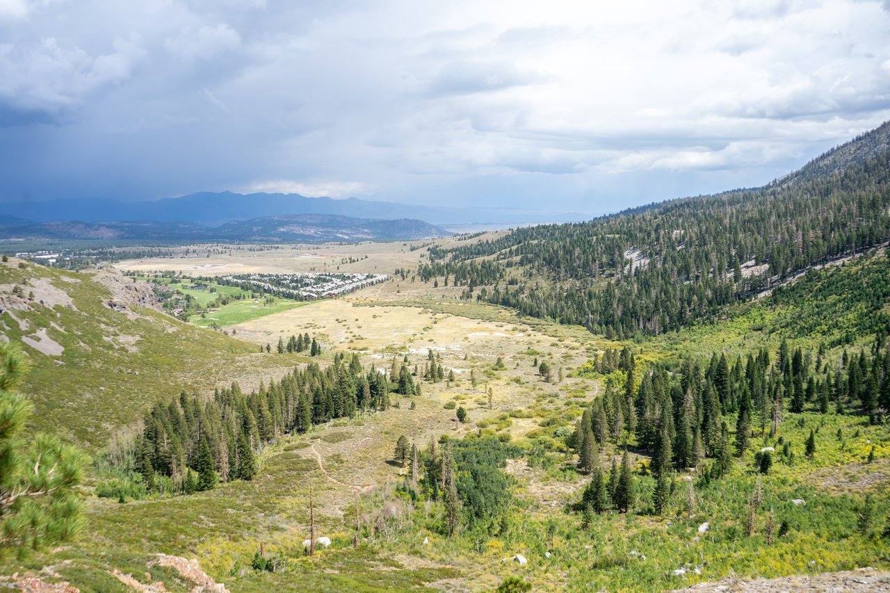 Mountain valley with pine-covered slopes, open meadows, and distant buildings beneath a cloudy sky.