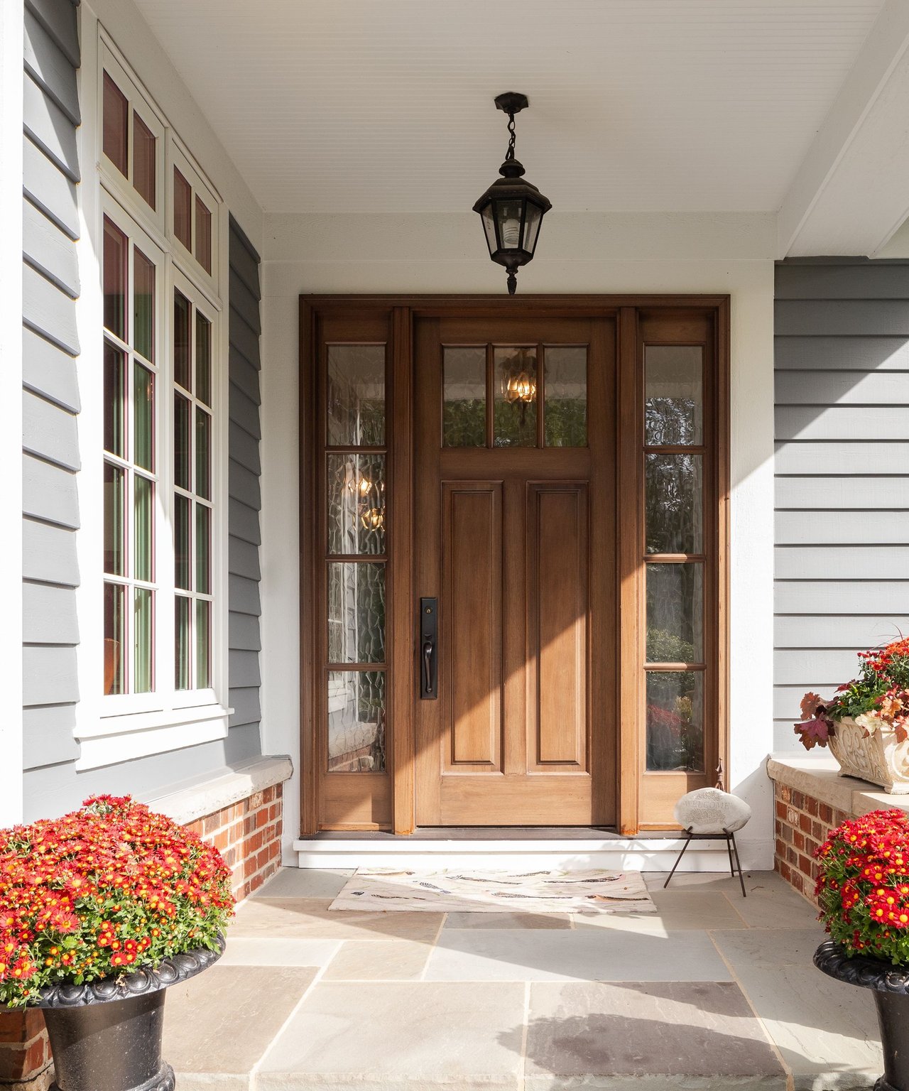 Covered front porch with a dark wood door, glass sidelights, stone paving, and pots of colorful flowers.