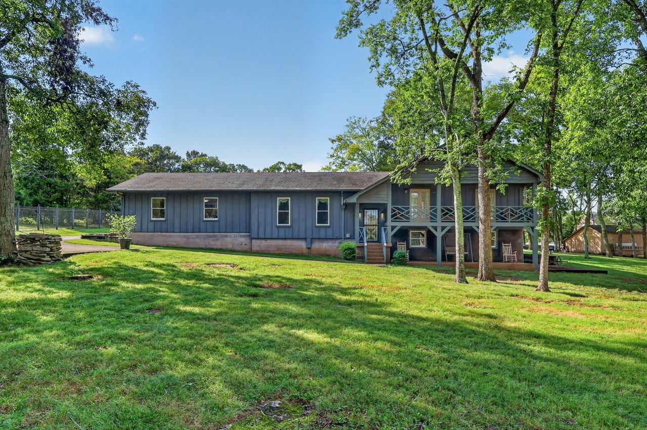 Blue home with a raised deck and covered porch beneath mature trees.