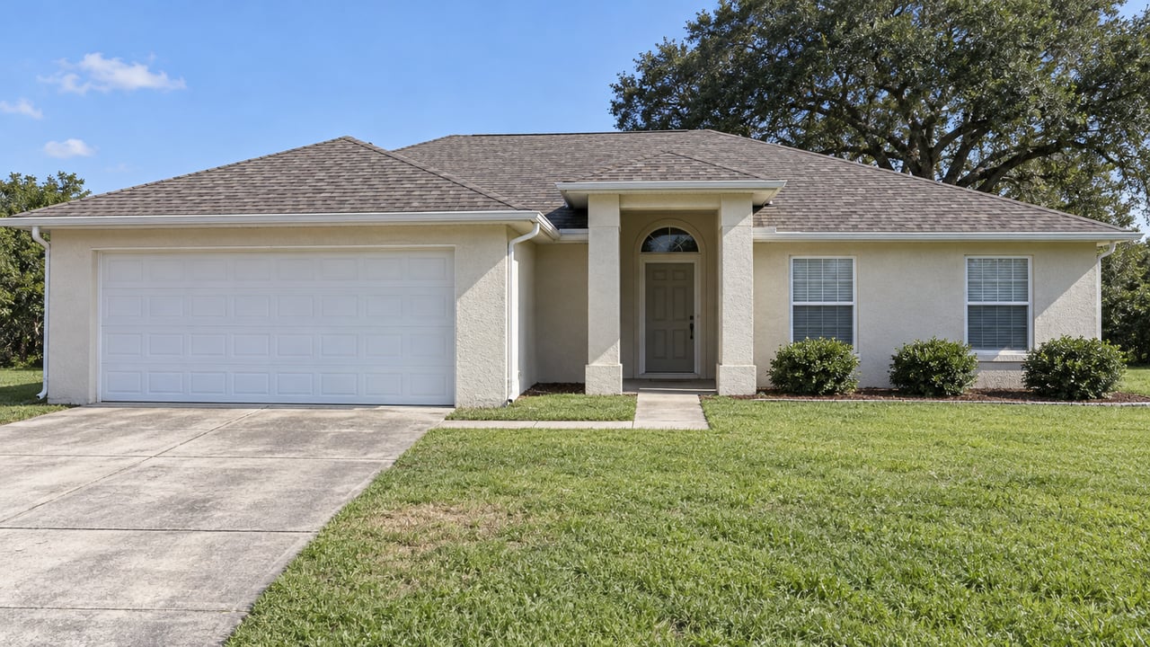 A pale stucco single-story home with a white garage, recessed entry, broad lawn and mature oak in bright daylight.