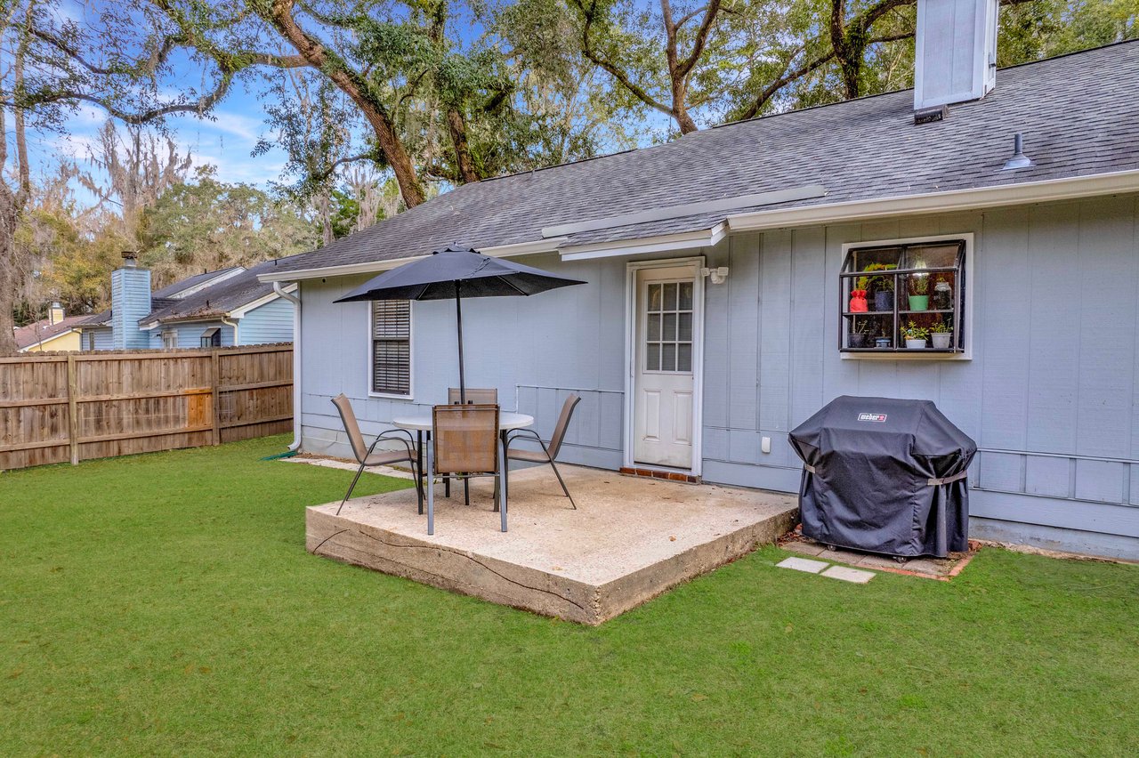 Backyard patio with a small table and chairs under a black umbrella. A covered barbecue grill is nearby, with potted plants on the window sill.