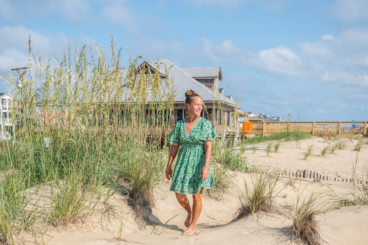 Amber Elicker standing barefoot among beach grass beside a coastal home.