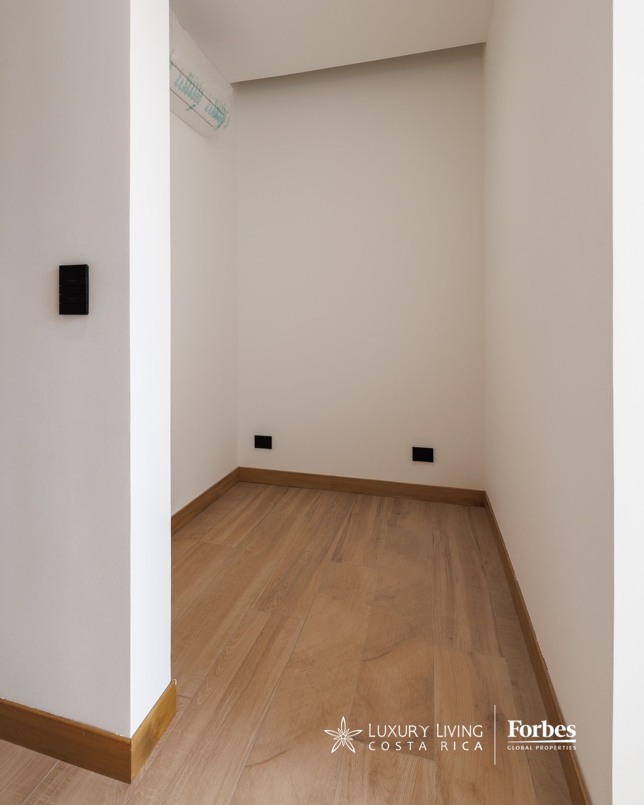 Empty narrow interior nook with wood-look flooring, wall outlets, and a high-mounted air conditioner.