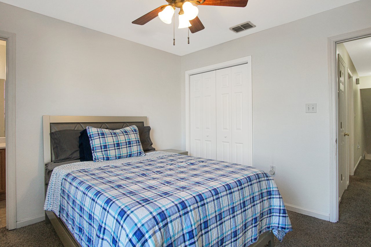 A bedroom with a cozy, inviting feel features a bed with a blue plaid comforter, gray pillows, light gray walls, and a ceiling fan with wooden blades.A bedroom with a cozy, inviting feel features a bed with a blue plaid comforter, gray pillows, light gray walls, and a ceiling fan with wooden blades.