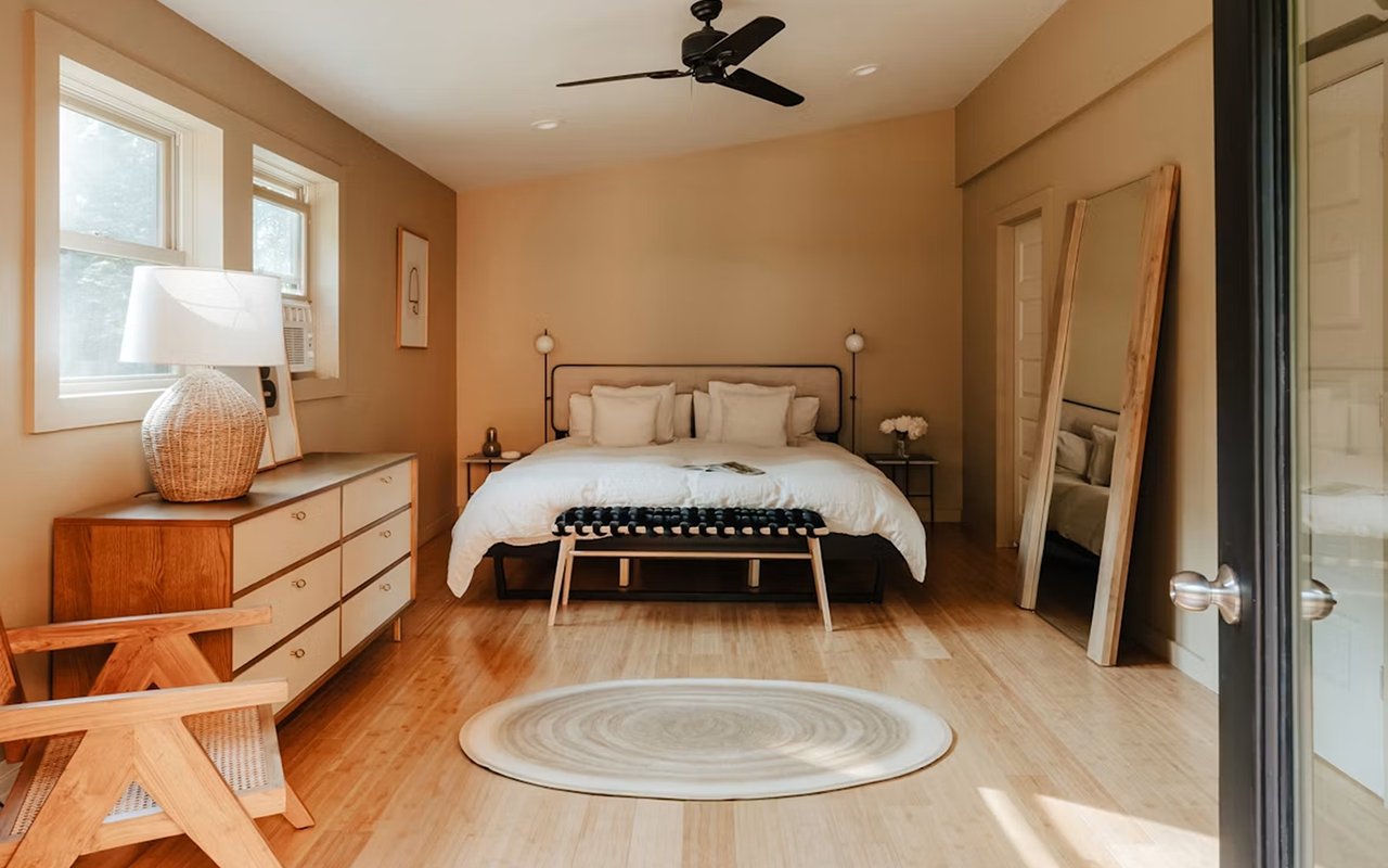 Furnished bedroom with a centered white bed, warm tan walls, wood floors, and a round rug.
