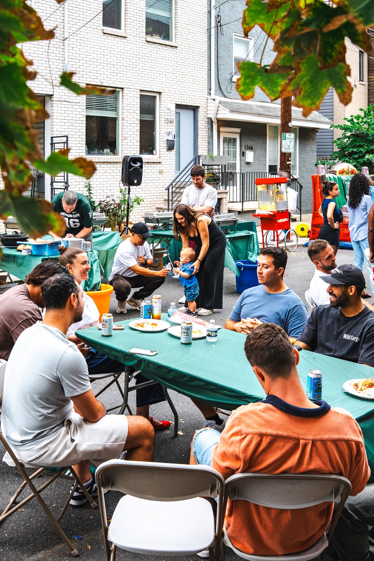 Neighbors share food at outdoor tables during a residential block party, with a child and popcorn machine nearby.
