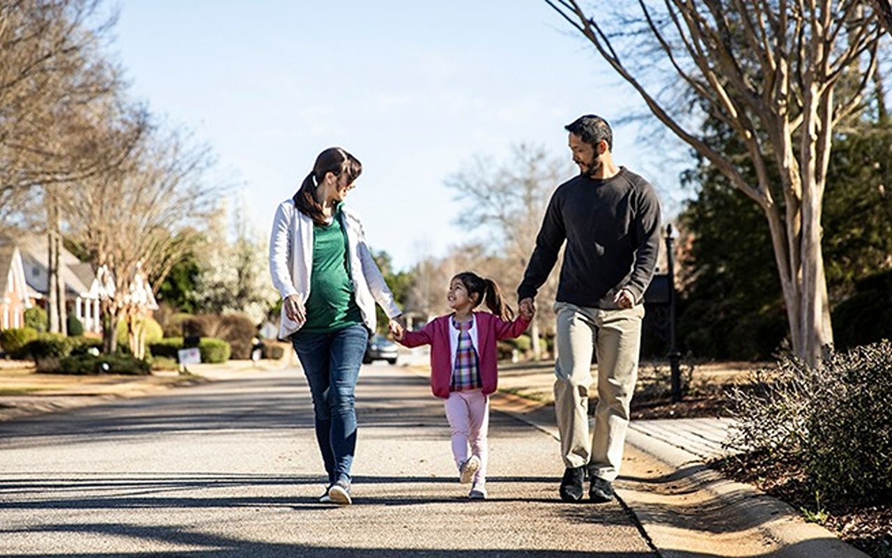 Couple walking with a young child on a sunny, tree-lined residential street.