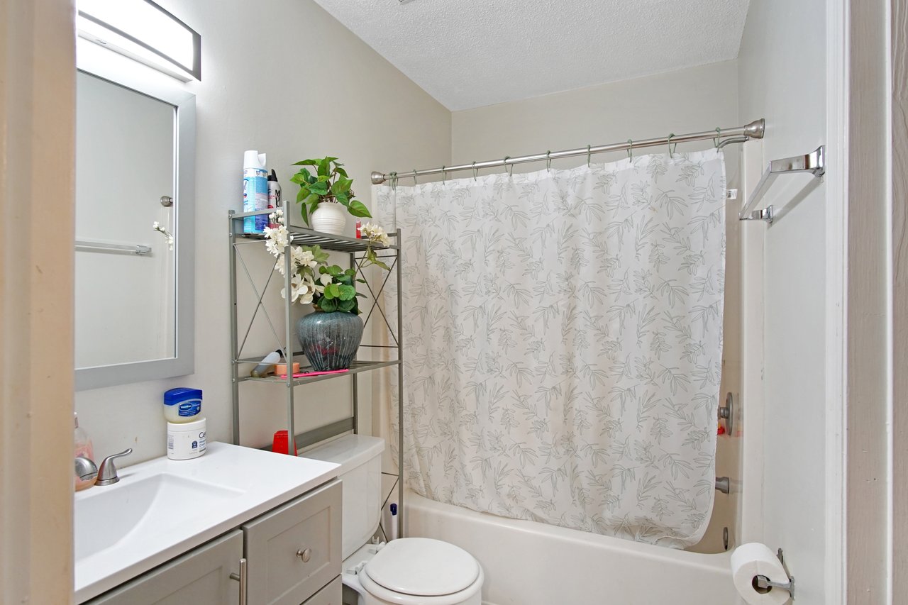 Bathroom at 2506 Nugget Lane in Tallahassee, Florida, with neutral walls and a white vanity topped with a modern faucet and mirror with overhead light. The space includes a toilet, metal shelving unit for storage, and a shower-tub combination with patterned curtain.