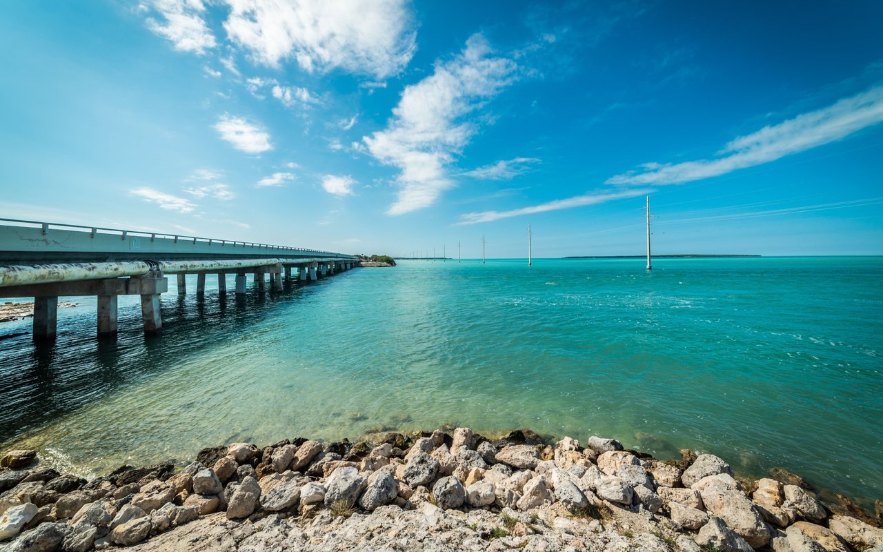 Bridge over turquoise water beside a rocky shoreline under a bright, cloud-streaked sky.