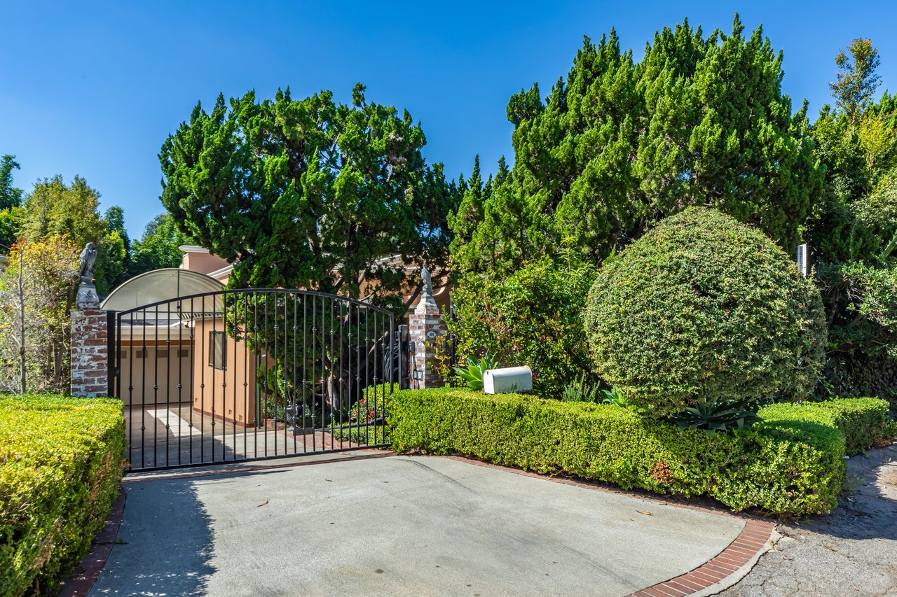 Gated residence entrance with clipped hedges, mature trees and a paved driveway.