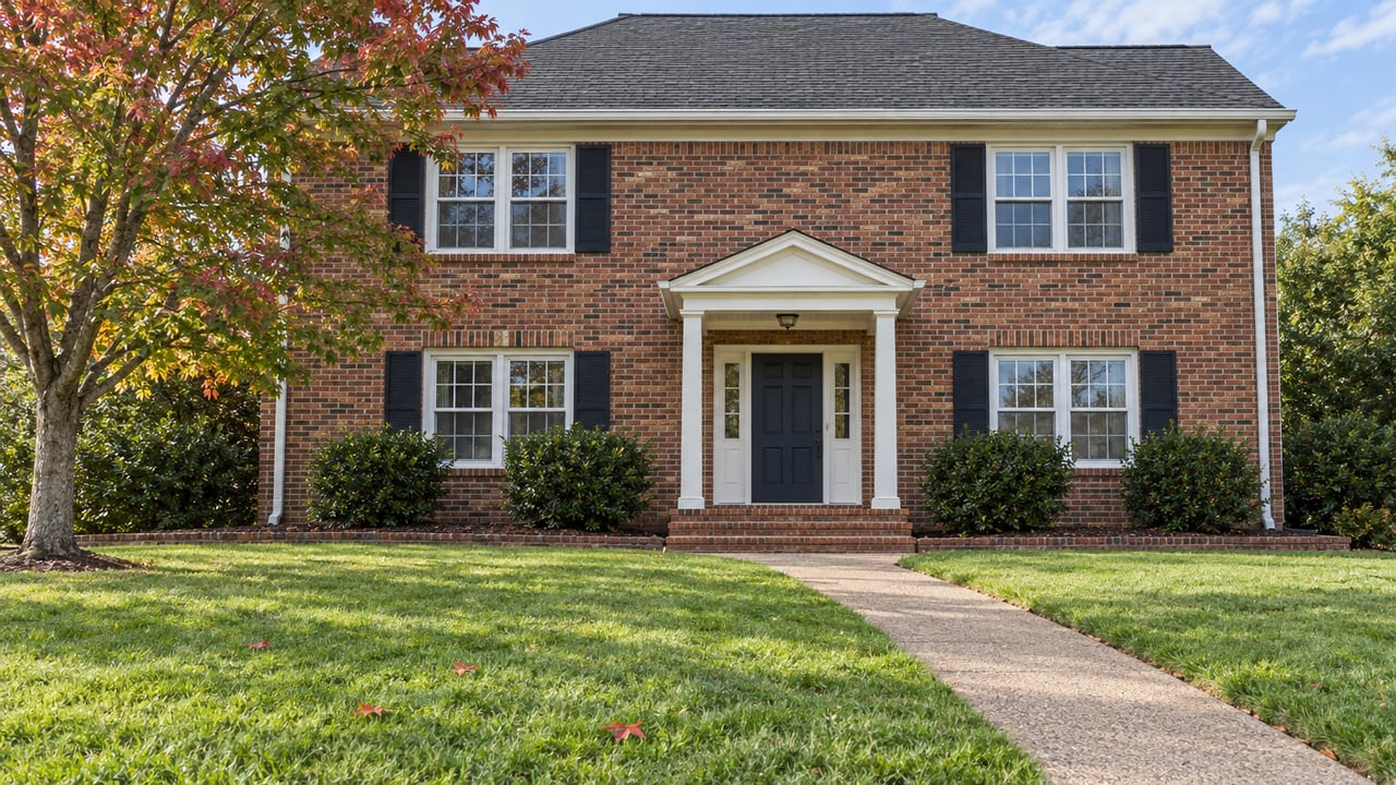 Two-story red-brown brick home with white window trim, black shutters and a small white portico beyond a broad autumn lawn.