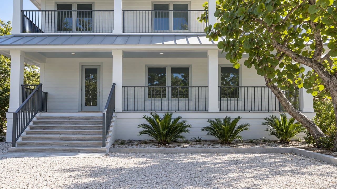 Sunlit white raised home with a pale metal roof, weathered timber entry stairs and a sea-grape tree beside its covered porch.