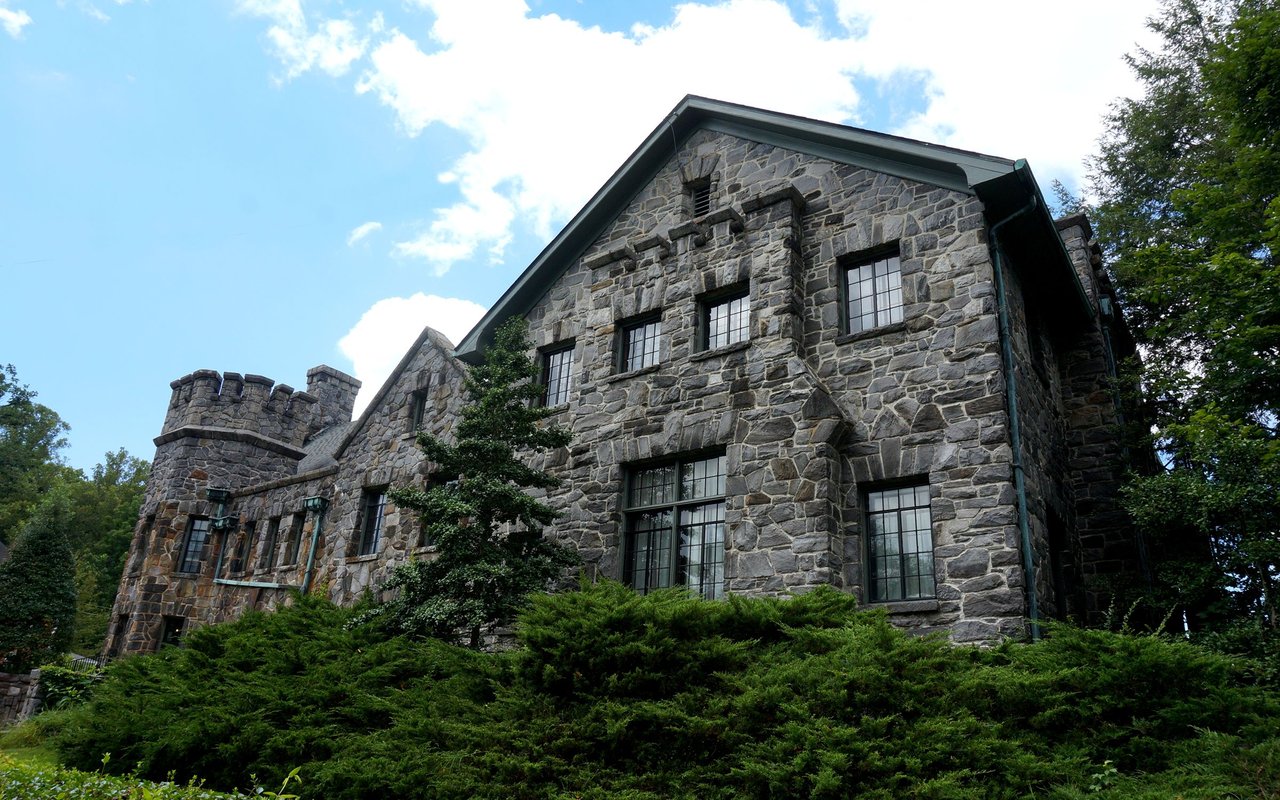 Stone house with a turret-like corner and tall gridded windows, framed by mature trees.