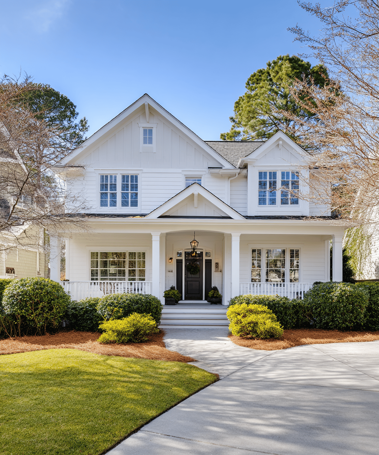 White two-story house with gabled roof, covered porch, landscaped shrubs, and concrete driveway.