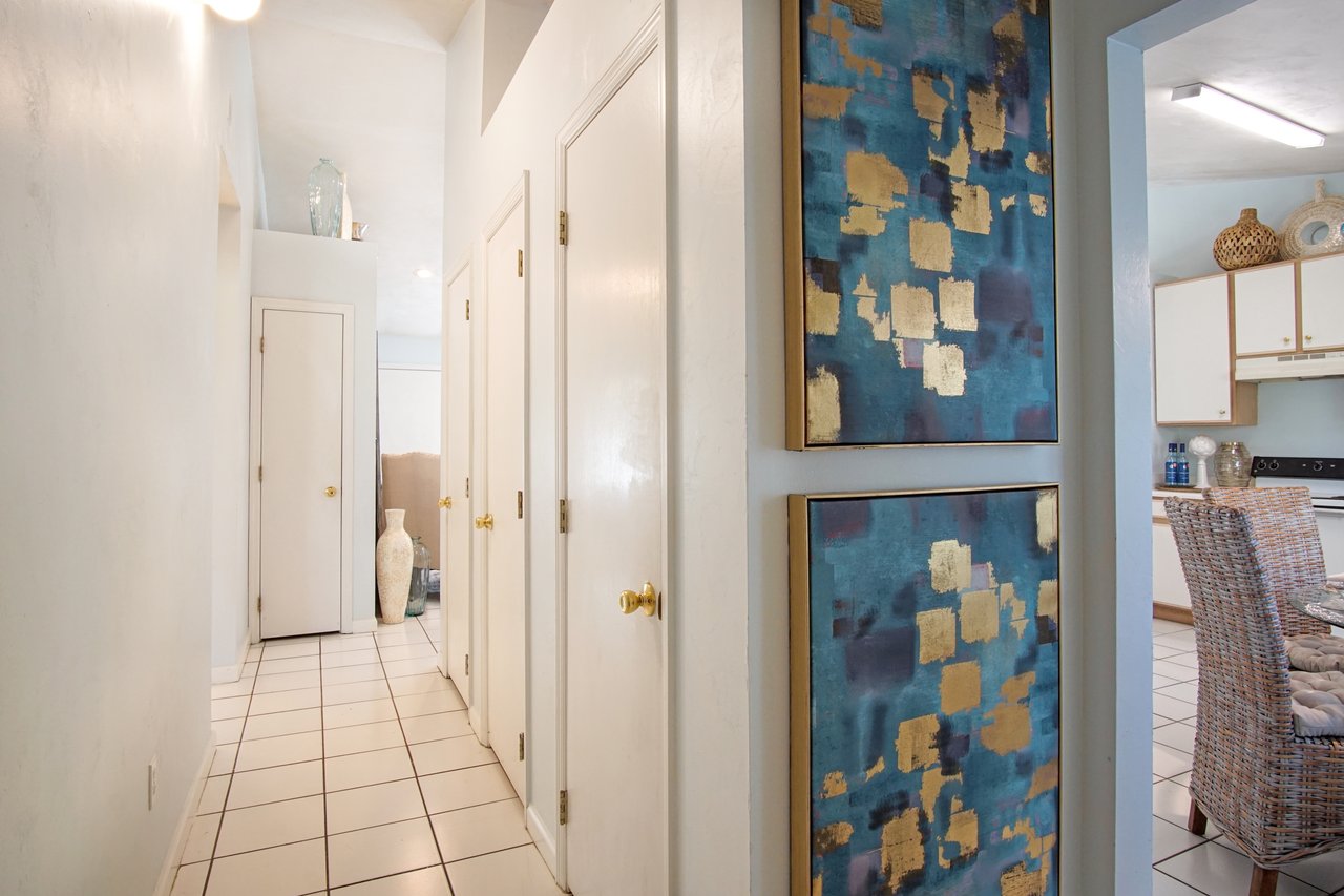 Narrow hallway with white tiled floor leading to a kitchen. Blue and gold abstract paintings decorate the wall. The vibe is modern and serene.