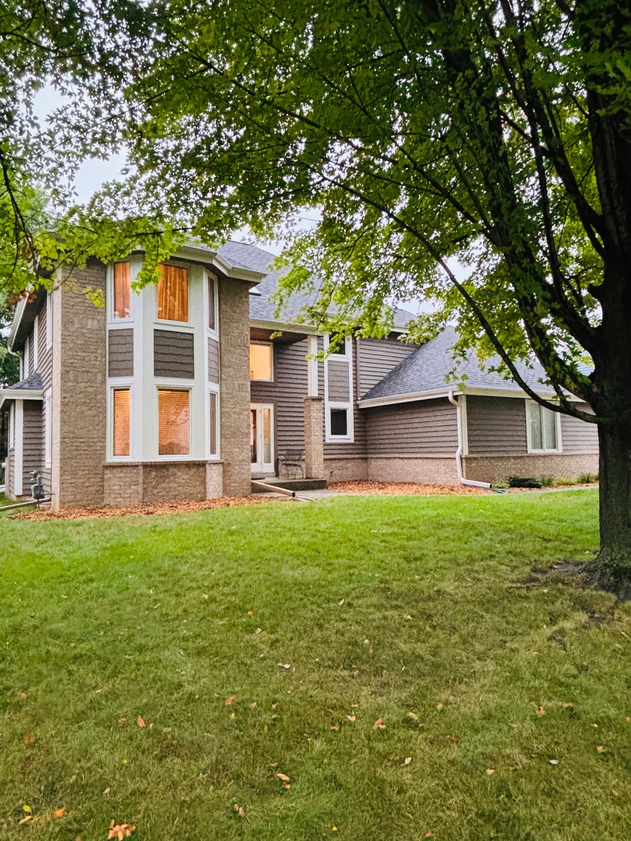Two-story stone and gray-sided house with bay windows beneath mature trees and a broad lawn.