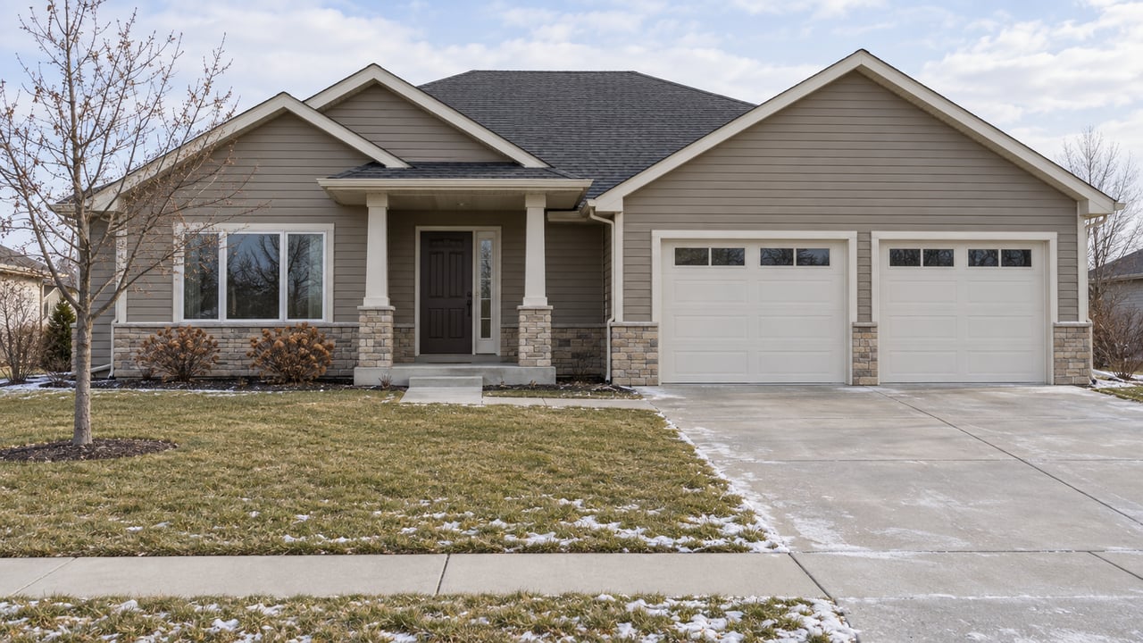 Taupe gabled home with twin garage doors, a covered stone-accented entry and a deep front lawn edged with light snow.